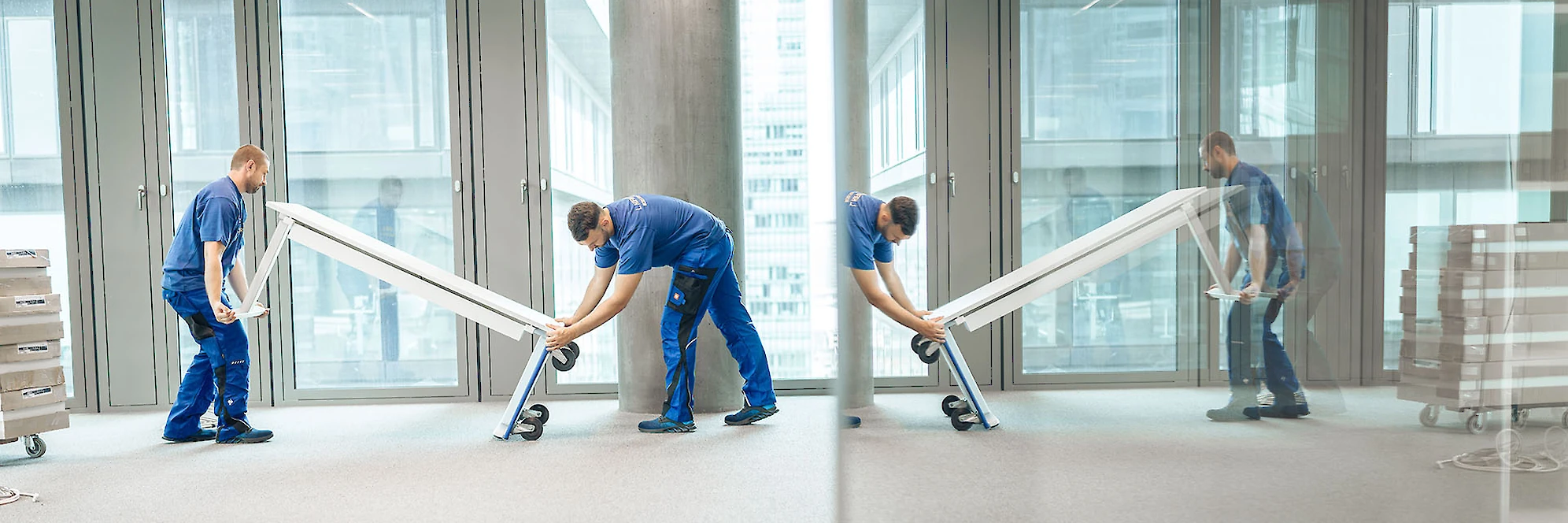 Zwei Mitarbeiter transportieren einen Tisch in einem modernen Bürogebäude mit großen Glasfenstern.