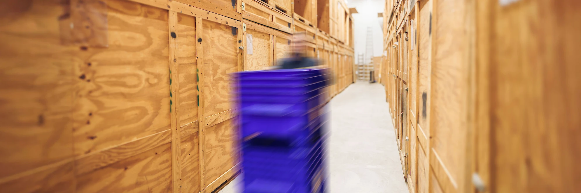 Dynamische Aufnahme eines Mitarbeiters in einem Lager mit Holzboxen, der eine blaue Transportkiste in einer Gasse zwischen den Lagerboxen bewegt.