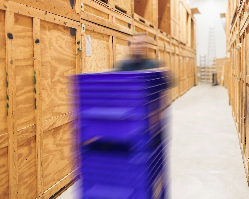 Dynamische Aufnahme eines Mitarbeiters in einem Lager mit Holzboxen, der eine blaue Transportkiste in einer Gasse zwischen den Lagerboxen bewegt.