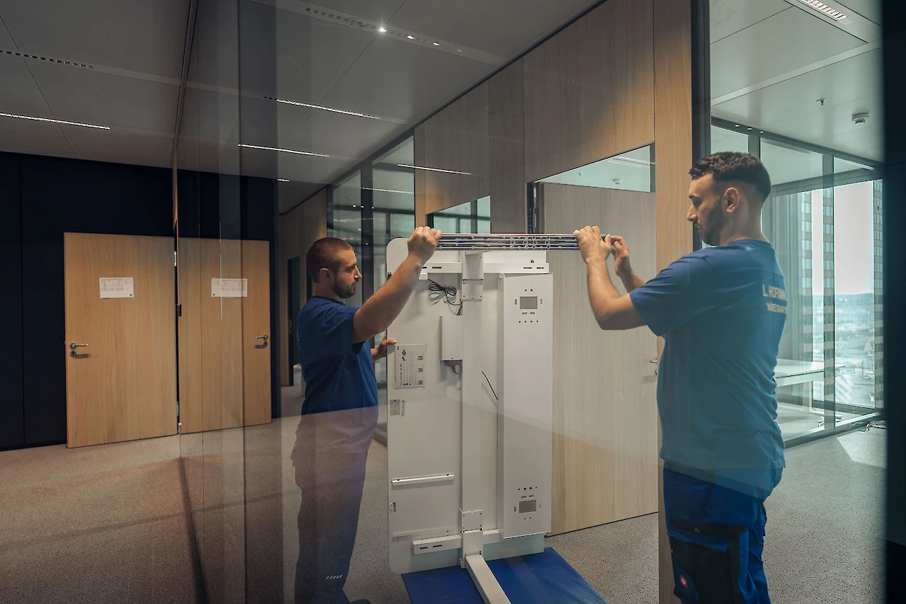 Zwei Mitarbeiter hantieren mit einem Gegenstand in einem Büroraum mit moderner Glas- und Holzeinrichtung.