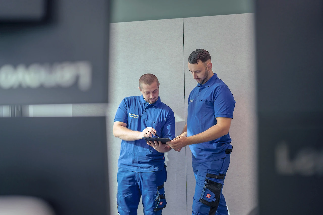 wei Mitarbeiter in blauer Arbeitskleidung überprüfen Details auf einem Tablet in einem modernen Lagerbereich.”