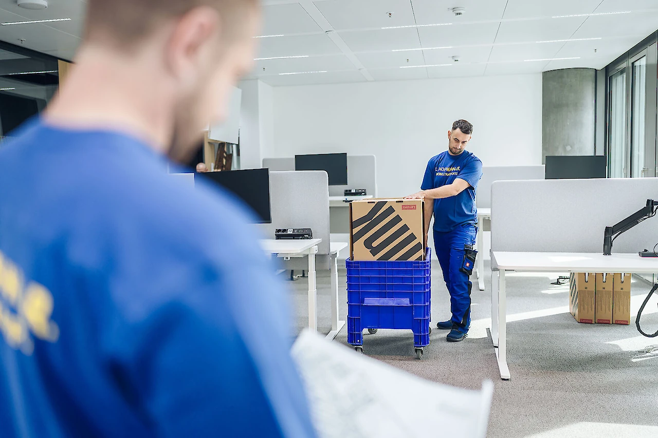 Mitarbeiter packt eine Box in einen blauen Rollcontainer, während ein Kollege einen Plan im Vordergrund prüft.