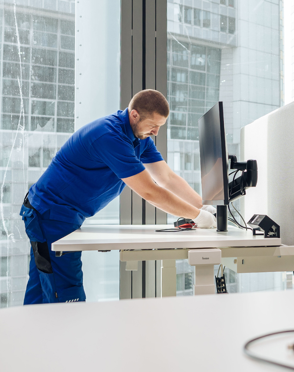 Techniker justiert präzise einen Monitor auf einem höhenverstellbaren Schreibtisch in einem modernen Büro mit Glasfassade.