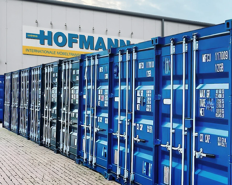 Reihe von blauen Containern vor einem Gebäude mit der Aufschrift ‘Hofmann Internationale Möbeltransporte’ für professionelle Zwischenlagerung.