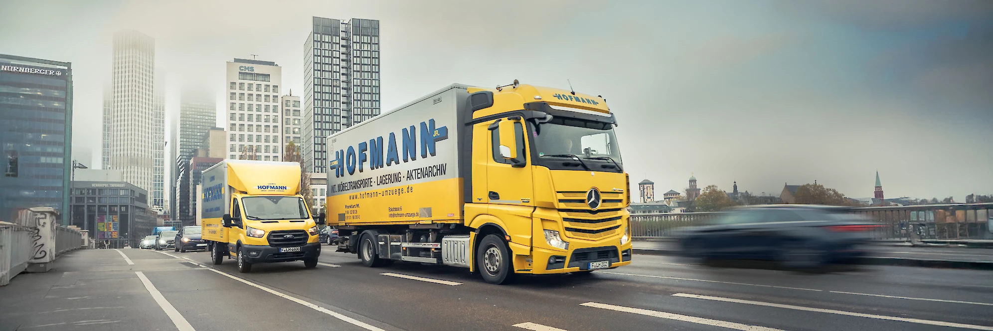 Zwei Hofmann Möbeltransporter fahren nebeneinander auf der Straße, im Hintergrund die Frankfurter Skyline.