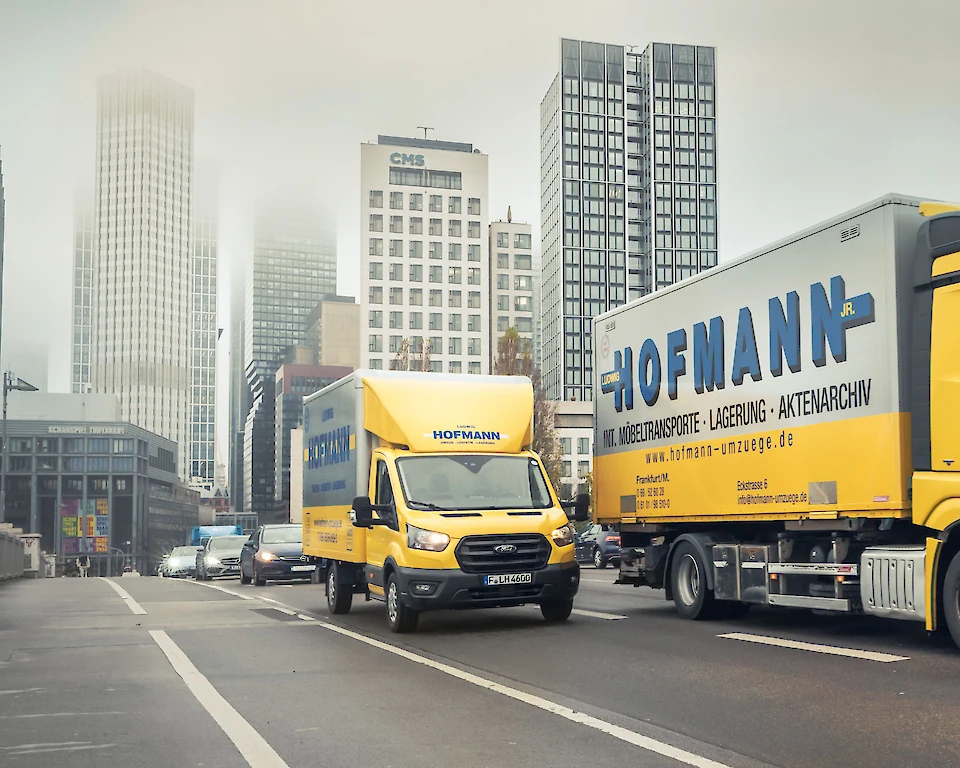 Zwei Hofmann Möbeltransporter fahren nebeneinander auf der Straße, im Hintergrund die Frankfurter Skyline.